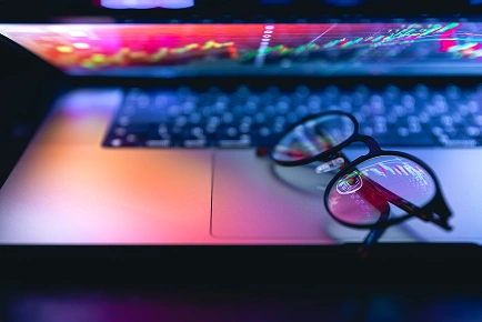 Óculos de grau sobre o teclado de um laptop aberto, com gráficos financeiros e de ações refletidos nas lentes.